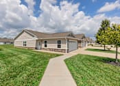 Thumbnail 2 of 21 - A house with a brown roof and a grey garage door is surrounded by a grassy lawn and a sidewalk.