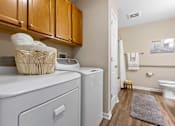 Thumbnail 15 of 21 - A laundry room with a washer and dryer, a basket of towels, and a rug on the floor.