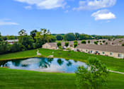 Thumbnail 19 of 20 - A serene landscape with a pond, houses, and trees under a clear blue sky.