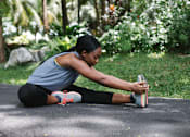 Thumbnail 12 of 12 - A woman stretches her leg while sitting on a mat outdoors.