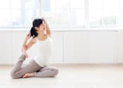 Thumbnail 10 of 12 - A woman in a white top and grey pants is sitting on the floor in a yoga pose.