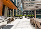 Thumbnail 20 of 56 - A patio with wooden furniture and a grill is surrounded by buildings.