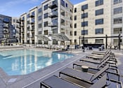 Thumbnail 9 of 107 - A large pool surrounded by sun loungers and umbrellas in front of apartment buildings.