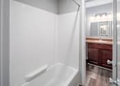 Thumbnail 24 of 25 - A white bathroom with a wooden vanity and a white bathtub.