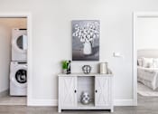 Thumbnail 23 of 37 - A white laundry room with a washer and dryer, a cabinet with a vase and plant on top, and a painting of flowers on the wall.