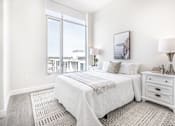 Thumbnail 26 of 37 - A bedroom with a large bed, a nightstand, and a window overlooking a cityscape.
