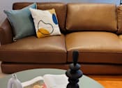 Thumbnail 9 of 10 - a round glass table with a mirror on top of it next to a brown leather couch