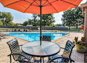Thumbnail 2 of 9 - A table with chairs and an umbrella is set up by a pool.