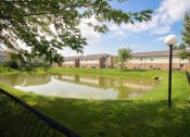 Thumbnail 8 of 14 - Pond in the middle of a grassy area with apartment buildings in the background at Maple Tree Apartments in LaPorte, IN
