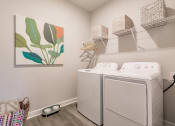 Thumbnail 26 of 57 - a laundry room with a washer and dryer and a painting on the wall at  a living room with tables and chairs and a ceiling fan at Swiftwater apartments in Port Charlotte, FL