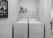 Thumbnail 22 of 51 - A laundry room with two washing machines and a shelf with laundry products.