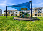 Thumbnail 7 of 45 - A playground with a blue canopy and a green slide is surrounded by apartment buildings.