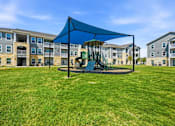 Thumbnail 5 of 45 - A playground with a blue canopy and a green slide is in front of apartment buildings.