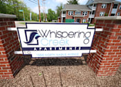 Thumbnail 1 of 34 - A sign for Whispering Creek Apartments is displayed in front of a brick wall.