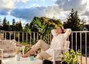 Thumbnail 5 of 10 - A person is sitting on a chair with their hands behind their head on a balcony.