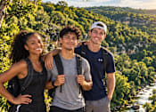 Thumbnail 112 of 128 - Three people standing on a rocky outcrop with a river and trees in the background.