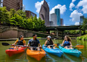 Thumbnail 48 of 52 - Four people kayaking on a river with a city skyline in the background.