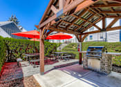 Thumbnail 18 of 34 - Happy Valley Apartments for Rent - Sunnyside Village - Outdoor Grilling Station Under a Large Built-In Awning with a Covered Stainless Steel Grill and Two Picnic Tables with Built-In Seating and Umbrellas