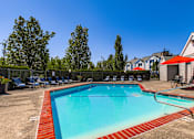 Thumbnail 15 of 34 - Happy Valley, OR, Apartments - Sunnyside Village - an Outdoor Gated Pool Area with Lounge Seating, Tables with Chairs and Umbrellas, Surrounding Trees and Shrubbery, and a View of the Property Building