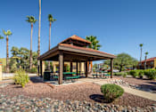 Thumbnail 6 of 13 - A gazebo with a red roof is surrounded by palm trees and a gravel area with a bench.