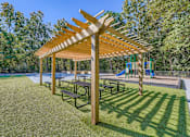Thumbnail 28 of 37 - A wooden pergola with benches is situated in a grassy area with a playground in the background.