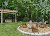 Thumbnail 27 of 39 - A wooden bench and chairs are arranged around a fire pit in a garden.