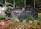 Thumbnail 24 of 24 - A sign for Bridge Creek Apartment Homes is surrounded by flowers and plants.