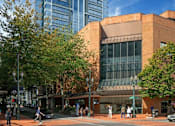 Thumbnail 77 of 78 - A modern glass building stands tall next to a smaller brick building with people walking around on the sidewalk.
