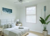 Thumbnail 13 of 14 - A bedroom with a bed, a ceiling fan, a nightstand with a lamp, a potted plant, and a painting on the wall.