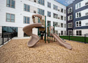 Thumbnail 18 of 19 - a playground with two slides and a chair in front of an apartment building at The Landing at Amber Fields Apartments, Rosemount, Minnesota