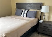 Thumbnail 13 of 28 - A bed with a white sheet and two pillows with a grey headboard and a nightstand with a lamp on it.