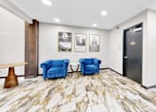 Thumbnail 16 of 34 - A hallway with a marble floor, two blue chairs, a wooden table, and pictures on the wall.