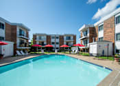 Thumbnail 1 of 12 - A swimming pool surrounded by red umbrellas and lounge chairs in front of apartment buildings.