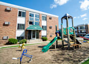 Thumbnail 9 of 12 - A playground with a green slide and a wooden swing set in front of a brick building with the number 90 on it.