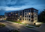 Thumbnail 69 of 92 - A modern apartment building is lit up at dusk with street lights on.