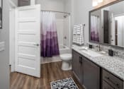 Thumbnail 19 of 24 - A bathroom with a white door, a purple shower curtain, and a grey granite countertop at The Edison at Woodbury, Woodbury, MN 55129