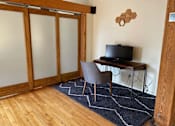 Thumbnail 9 of 20 - A room with a wooden floor, a chair, a table with a TV on it, and a rug.