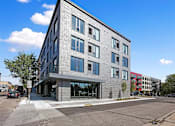 Thumbnail 44 of 54 - A modern building with a grey facade and large windows is situated on a street corner.