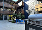 Thumbnail 7 of 9 - A silver grill is in the foreground of a modern building with blue chairs and tables.