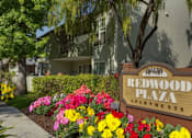 Thumbnail 1 of 8 - A sign for Redwood Plaza Apartments is in front of a building with a flower bed in the foreground.