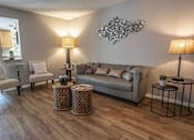 Thumbnail 8 of 10 - A living room with a grey couch, a chair, a table with a lamp and a plant on it, and a decorative piece on the wall.