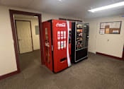 Thumbnail 6 of 6 - A Coca-Cola vending machine sits in a room with carpeted flooring and white walls.