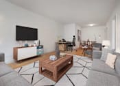 Thumbnail 2 of 12 - A living room with a grey couch, a wooden coffee table, and a television on the wall at The Caldwell Apartments, New York