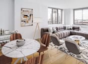 Thumbnail 10 of 17 - A modern living room with a large grey sofa, a round marble table, and a view of the city at The Ellington Apartments, New York, 10019