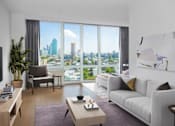 Thumbnail 4 of 15 - A modern living room with a view of the cityscape outside the large window at The Maximilian Apartments, Long Island City, NY