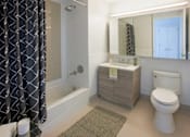 Thumbnail 14 of 15 - A bathroom with a white toilet, a sink, and a bathtub with a grey shower curtain at The Maximilian Apartments, Long Island City, New York