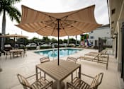 Thumbnail 14 of 23 - A large umbrella shades a table and chairs by a pool.