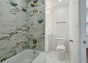 Thumbnail 12 of 34 - A white toilet sits in a bathroom with a glass mosaic wall at Shipyard Shores Apartments, Newport News