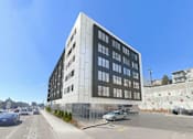 Thumbnail 9 of 22 - A modern building with a glass facade is situated on a street with cars and a pedestrian crossing.