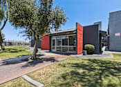 Thumbnail 11 of 42 - A modern building with a red and grey exterior is surrounded by a grassy area and a tree.